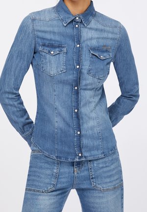 Woman wearing a fitted, long-sleeve denim shirt with buttoned chest pockets and matching denim jeans.