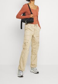 Rippiges rostfarbenes Langarmoberteil kombiniert mit beigen Cargo-Hosen, die ein strukturiertes, wirbelndes Muster aufweisen. Die Schuhe sind silberne Sneakers und eine schwarze Tasche.