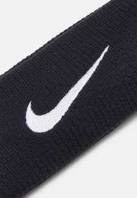 Un bandeau de sport noir en tissu côtelé avec un logo Nike Swoosh blanc bien visible. Surface texturée pour une meilleure adhérence et confort.
