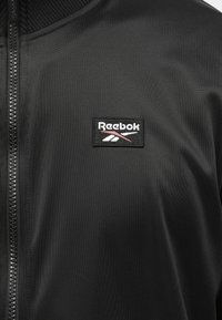 Artimai nufotografuota juoda Reebok striukė su priekiniu užtrauktuku ir mažą stačiakampį Reebok logotipą kairėje krūtinės pusėje.