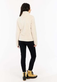 Fleece jack in crème met een hoge kraag, gecombineerd met zwarte leggings en tan laarzen met zwarte accenten en bontvoering.