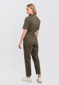 Olive green jumpsuit with short sleeves, fitted waist, and tapered legs. Made of smooth fabric, featuring a collar and rear pockets.