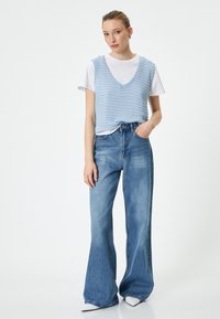 Chaleco de punto azul claro sobre una camiseta blanca, combinado con unos vaqueros de campana azul claro de cintura alta. La modelo está de pie frente a un fondo simple.
