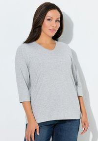 Top grigio chiaro con scollo a V e maniche a tre quarti, realizzato in tessuto morbido, che presenta una vestibilità comoda. La texture è liscia con lievi variazioni.