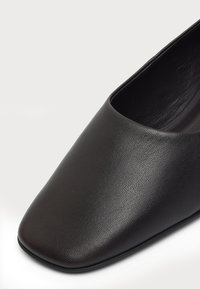 Gros plan d'une chaussure en cuir noire avec un bout carré et une surface lisse texturée sur fond blanc.