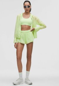 Neon zöld sportöltözet, amely tartalmaz egy crop topot, rövidnadrágot és egy könnyű dzsekit. Fehér sportcipő és csíkos zoknik egészítik ki, a stílusos megjelenés érdekében.