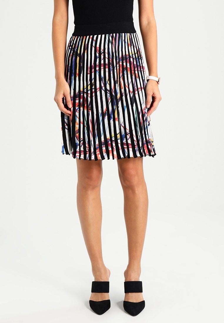 Femme portant une jupe plissée colorée avec des rayures verticales noires, un top noir, des talons noirs à brides et une montre blanche au poignet.