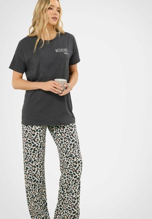 Vrouw die een mok vasthoudt, draagt een donker T-shirt met de tekst "Weekend Vibes" en een broek met luipaardprint, staande tegen een witte achtergrond.