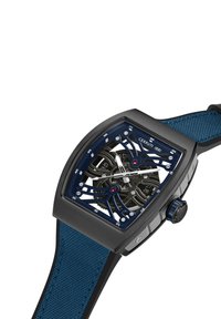 Cerruti-klocka med fyrkantig svart boett, blå skelettskiva, silverfärgade visare och ett strukturerat blått textilarmband. Accenter inkluderar en röd detalj.