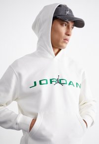 Fehér kapucnis pulóver elülső zsebbel és zöld "JORDAN" logóval, fekete sziluettel egy játékosról. Szürke sapka megkopott részletekkel.