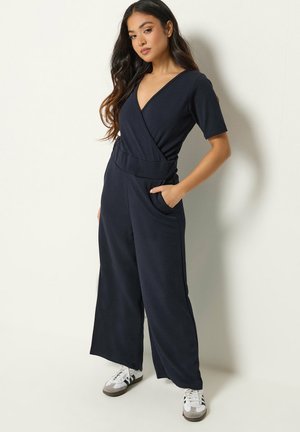 Kvinde med langt mørkt hår iført en sort slå-om-jumpsuit og hvide sneakers, stående med den ene hånd i lommen mod en ensfarvet væg.