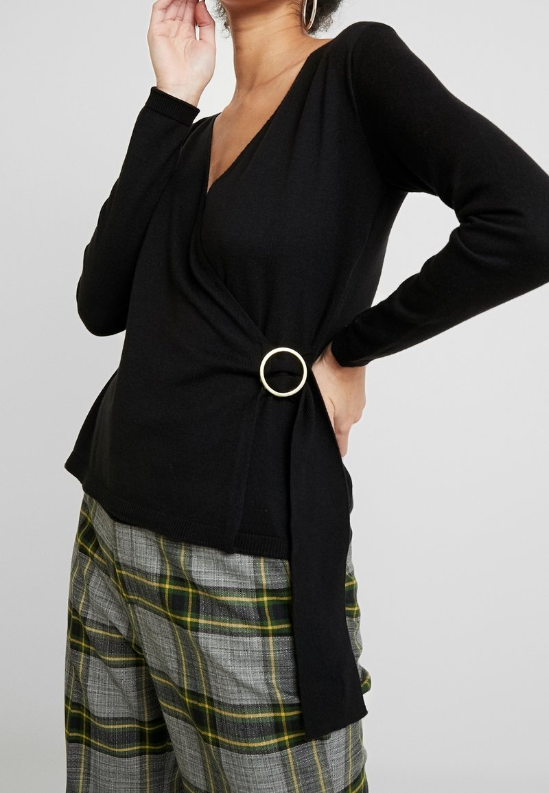 Černý top s rukávy ve měkké pletenině s výstřihem do V a zlatým kroužkem na boku, kombinovaný s kostkovanými kalhotami v šedé a zelené.
