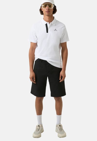 White polo shirt with a black placket and logo, paired with black shorts featuring zip pockets. Model wears white socks and beige sneakers.