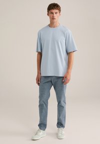 Helltürkises Baumwoll-T-Shirt mit kurzen Ärmeln, kombiniert mit strukturierten blauen Hosen und weißen Turnschuhen, die vor einem neutralen Hintergrund stehen.