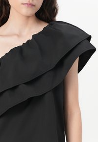 Svart topp med en fremtredende volangdetalj på skulderen, med en glatt tekstur og asymmetrisk design som skaper en dristig silhuett.