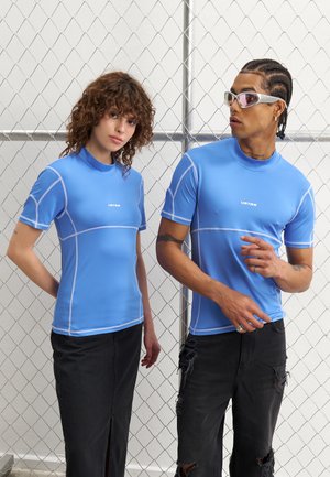 Zwei Models tragen figurbetonte, hellblaue kurzärmlige Hemden aus glattem Material, die kontrastierende weiße Nähte und ein Logo in der Mitte aufweisen.