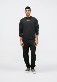 Μαύρη μπλούζα με στρογγυλή λαιμόκοψη και λογότυπο στο στήθος, συνδυασμένη με μαύρες φόρμες. Το υλικό φαίνεται απαλό με άνετη εφαρμογή. Γκρίζα αθλητικά παπούτσια.