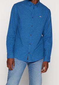 Blaues Hemd mit Knöpfen und strukturiertem Muster, ausgestattet mit einem Kragen und dunklen Knöpfen. Kombiniert mit hellblauen Jeans.
