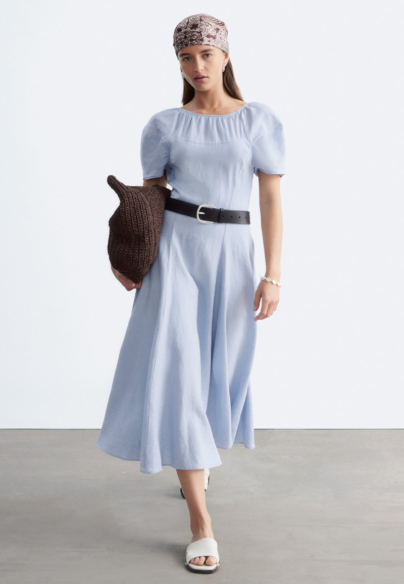 Femme marchant portant une robe bleu clair à manches bouffantes, ceinture noire, sandales blanches, foulard à motifs, tenant un sac en tricot marron sur fond uni.