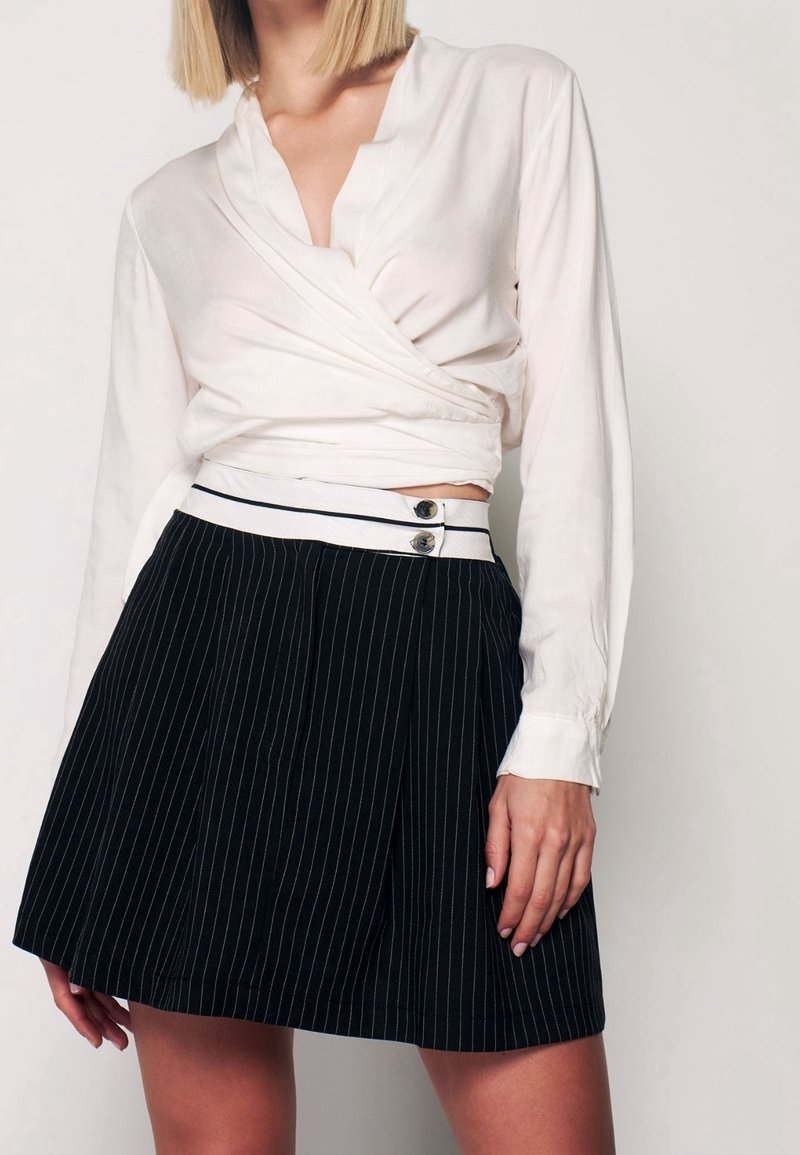 Femme portant un chemisier blanc style cache-cœur et une jupe noire à fines rayures taille haute avec une ceinture à double bouton, debout devant un fond uni.