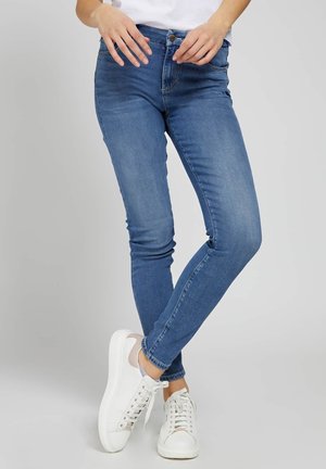 Vrouw draagt blauw slim-fit spijkerbroek en witte sneakers, staand met gekruiste benen tegen een effen achtergrond.