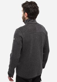 Veste en polaire grise avec un col haut, tissu texturé, couture visible et écusson logo sur le bras gauche. Coupe classique, manches longues.