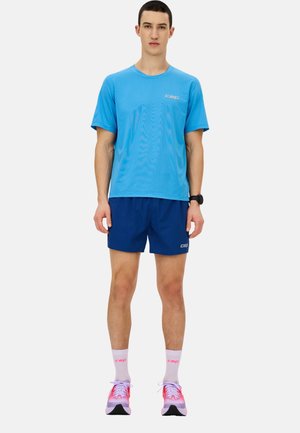 Blaues Sport-T-Shirt, marineblaue Shorts, rosa und schwarze Laufschuhe mit weißen Socken. Das T-Shirt hat ein Logo auf der Brust.