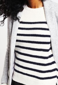 Cardigan grigio sopra un maglione bianco con strisce orizzontali blu navy. Il maglione presenta una texture a coste, mentre il cardigan mostra un tessuto liscio.