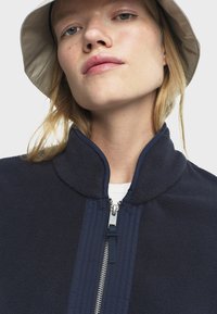 Chaqueta de forro polar de color azul marino con cuello alto, cierre frontal de cremallera y detalles de panel contrastante. El modelo lleva un sombrero de cubo beige.
