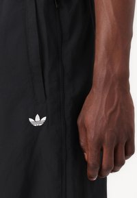 Svarta träningsshorts tillverkade av lättviktsmaterial, med en central dragkedja och en vit broderad logotyp på vänster sida.