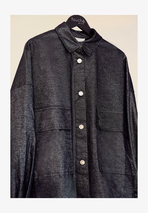 Fekete, fémes gombos ing, gallérral, patentos záródással és elülső zsebbel. Simább textúrájú és laza vágású dizájn.