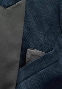 Dunkelblaues strukturiertes Gewebe mit geometrischen Mustern, akzentuiert durch ein glattes, dunkelgraues Taschentuch mit einem dezenten hexagonalen Design.