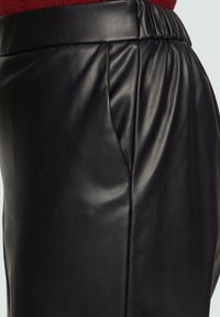 Zwarte leren broek met een geplooid ontwerp, een gladde textuur en een elastische tailleband. Focus op de zijaanzicht dat de kwaliteit van het materiaal toont.