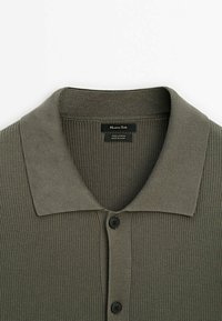 Cotton polo shirt in muted olive green. Features a ribbed texture, flat collar, and three black buttons down the front. Care label visible.