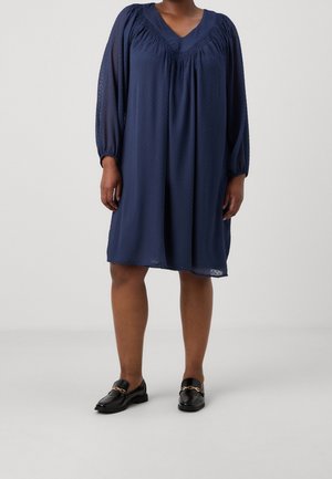 Frau trägt ein locker sitzendes dunkelblaues Langarmkleid mit gerafften Details und schwarze Loafers mit goldenen Kettendekorationen.