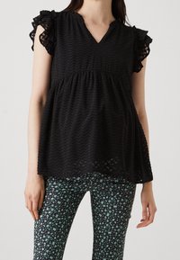 Blusa nera con scollatura a V, maniche a volant e motivo a zigzag testurizzato, abbinata a pantaloni neri con una piccola stampa floreale verde.