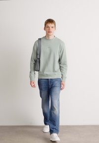 Felpa verde chiaro con collo rotondo, polsini e orlo a costine, abbinata a jeans blu e sneakers bianche. Il modello porta una borsa a tracolla grigia.