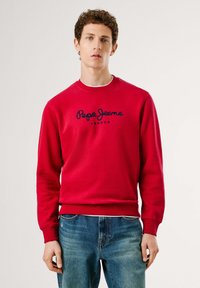 Joven vestido con sudadera roja de Pepe Jeans London y jeans azules, de pie frente a un fondo claro y liso.