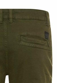 Olivgrüne Hose mit einer Gesäßtasche und einem rechteckigen Aufnäher mit Logo, auf dem Textur- und Nähdetails zu sehen sind.