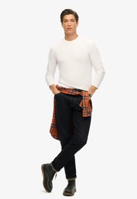 Witte lange mouwen shirt, donkerblauwe spijkerbroek, zwarte laarzen en een oranje geruit shirt om de taille geknoopt. Eenvoudig ontwerp zonder zichtbare logo's.