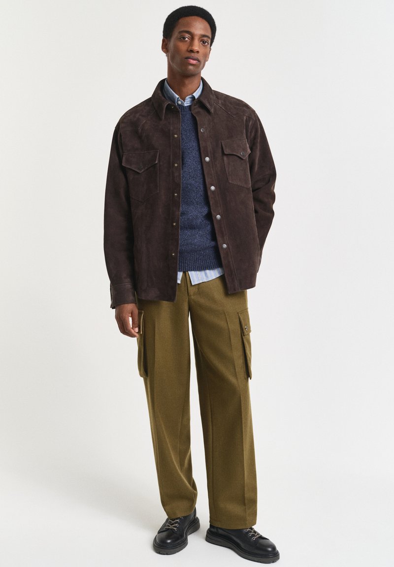 Uomo con giacca in suede marrone scuro, maglione blu sopra camicia a righe, pantaloni cargo oliva e scarpe nere, in piedi davanti a uno sfondo bianco.