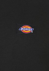 Camiseta de algodón negra con un parche rectangular de logo con borde rojo y amarillo y texto azul "Dickies". Textura suave, diseño ajustado.