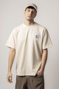Tricou din bumbac bej, cu mâneci scurte, guler rotund și un mic design grafic albastru pe pieptul stâng. Asortat cu pantaloni maro.