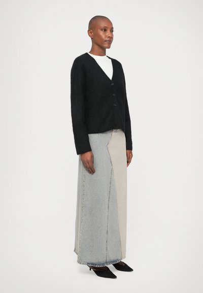 Cardigan preto de malha com decote em V e cinco botões, usado sobre uma saia de ganga longa em vários tons. O modelo está de saltos agudos pretos.