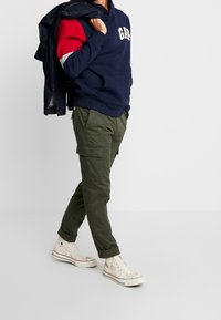 Felpa navy con logo "GAP", maniche con accenti rosso e bianco, pantaloni cargo oliva e sneakers bianche alta, giacca di jeans tesa sulla spalla.