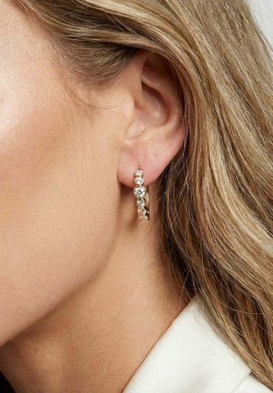 Guld ørering med syv runde klare ædelstene båret i en kvindes venstre øre, med blondt hår og lys hud synlig.