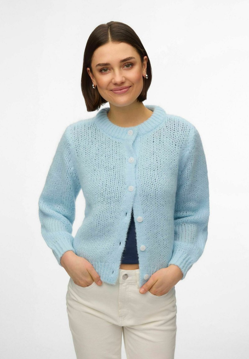 Kvinne med kort mørkt hår som har på seg en lyseblå strikket cardigan over en marineblå topp og hvite bukser, smilende med hendene i lommene.