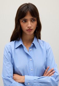 Giovane donna con lunghi capelli castani, indossa una camicia azzurra a righe con bottoni, braccia incrociate, guarda leggermente di lato con un'espressione neutra.