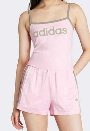 Kobieta w jasno różowej koszulce adidas z szarym wykończeniem i pasujących szortach z bocznymi paskami i logo, pozująca na jednolitym tle.