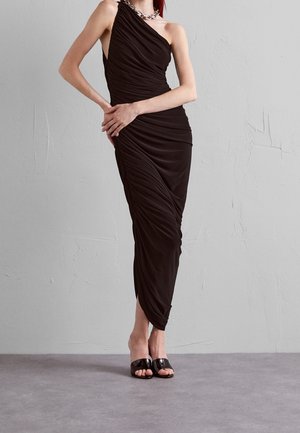 Vrouw gekleed in een gerimpelde, asymmetrische zwarte jurk met een zilveren ketting en zwarte open sandalen, staand tegen een grijze, gestructureerde muur.